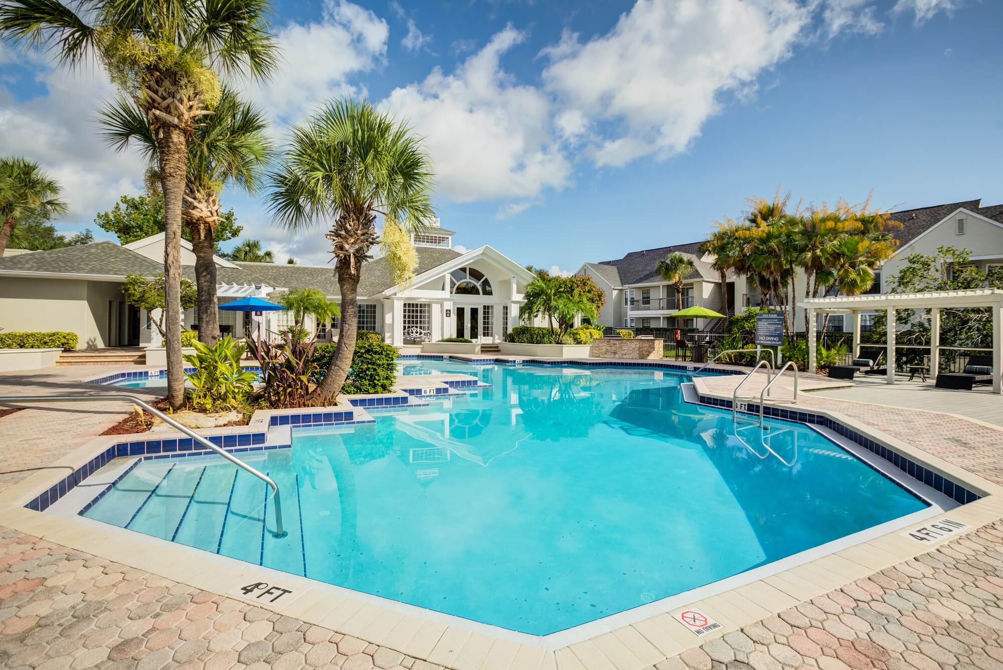 side view of large pool with palm trees and view of clubhouse and shaded lounge area.